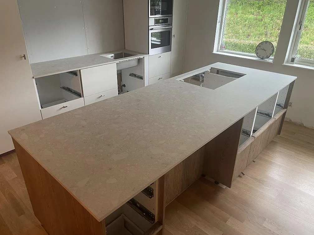 Køkkenbordplade og stor køkkenø i mat Dekton Ava, varmt cremefärgad keramik med terrazzomönster, med ekfronter og infälld induktionshäll i ett køkken under montering i Falun.