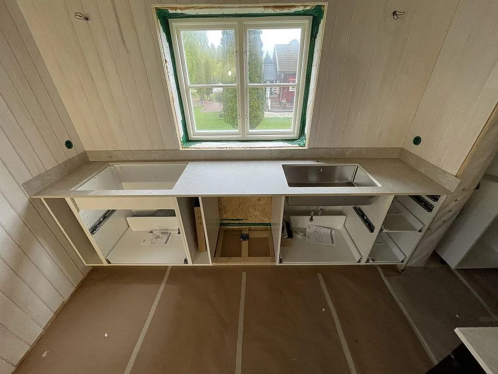Køkkenbordplade i mat Dekton Ava, lys grågrön keramik, med underlimmad rostfri vask under fönstret i ett køkken i Mora.