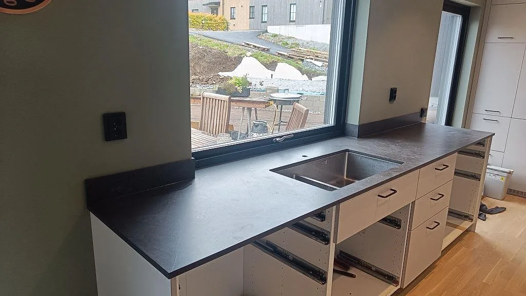 Køkkenbordplade i mørk mat Dekton Fossil med låg stänkkant og vask vid vindue i ett køkken i Landvetter.