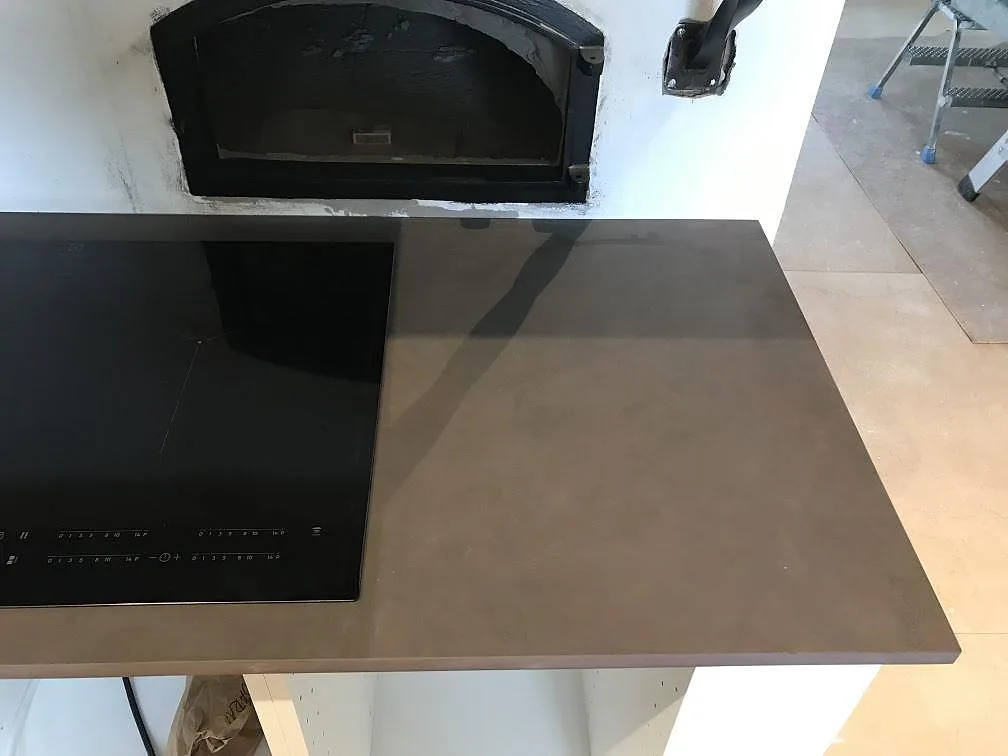 Køkkenbordplade i mörkt grågrå Dekton Keranium, mat, med infälld induktionshäll bredvid en murad vedeldad bakugn i ett køkken i Moheda.