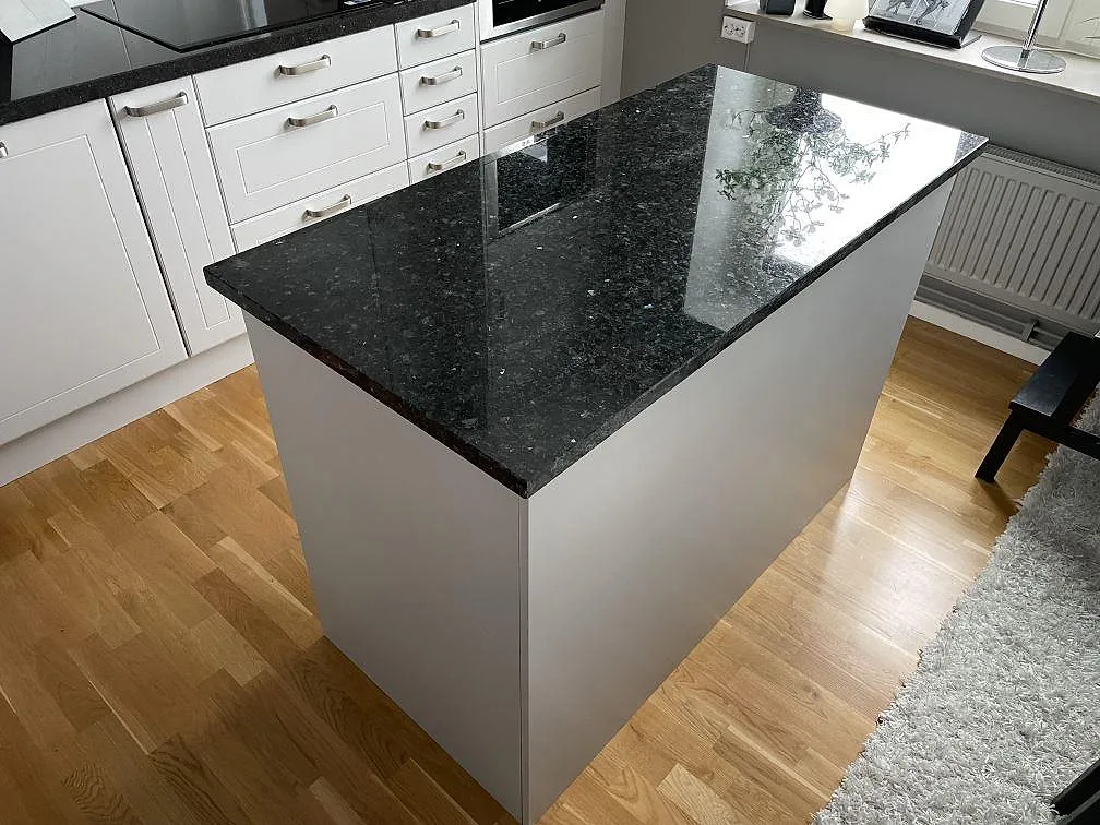 Køkkenø med poleret Bordplade i Emerald Pearl granit, mørk natursten med blågröna skiftningar, i ett vitt køkken i Tyreso.