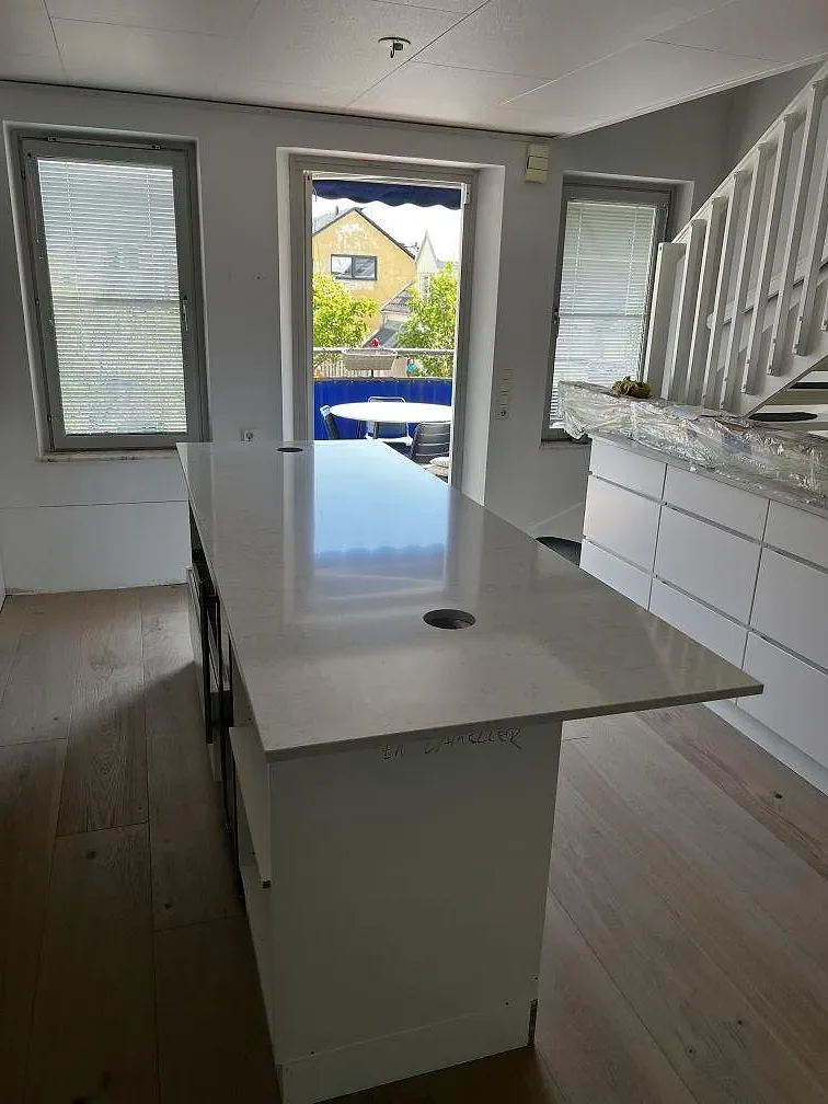 Køkkenbordplade i hvid Silestone Snowy Ibiza, poleret kvartskomposit, monteret som køkkenø med ljust ådrad overflade i ett køkken under montering i Limhamn.