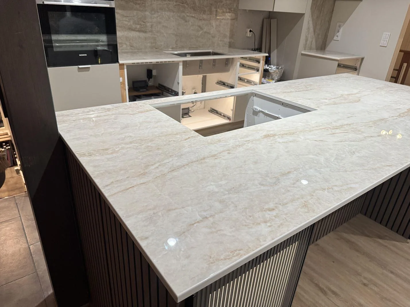 Køkkenbordplade i poleret Makrana keramik med varm beige åretegning, monteret som halvö med mørke räfflade fronter i ett køkken under montering i Markaryd.