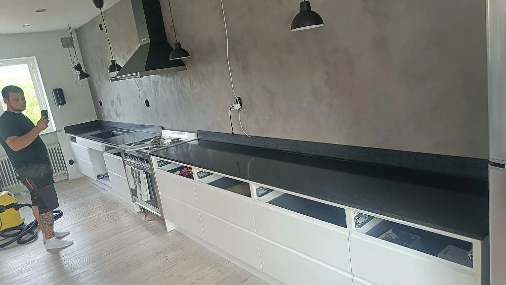 Køkkenbordplade i sort Nero Assoluto, mat granit, längs en lang enradsvägg med vita greppfria underskåp og sort fläktkåpa i ett køkken i Köpingebro.