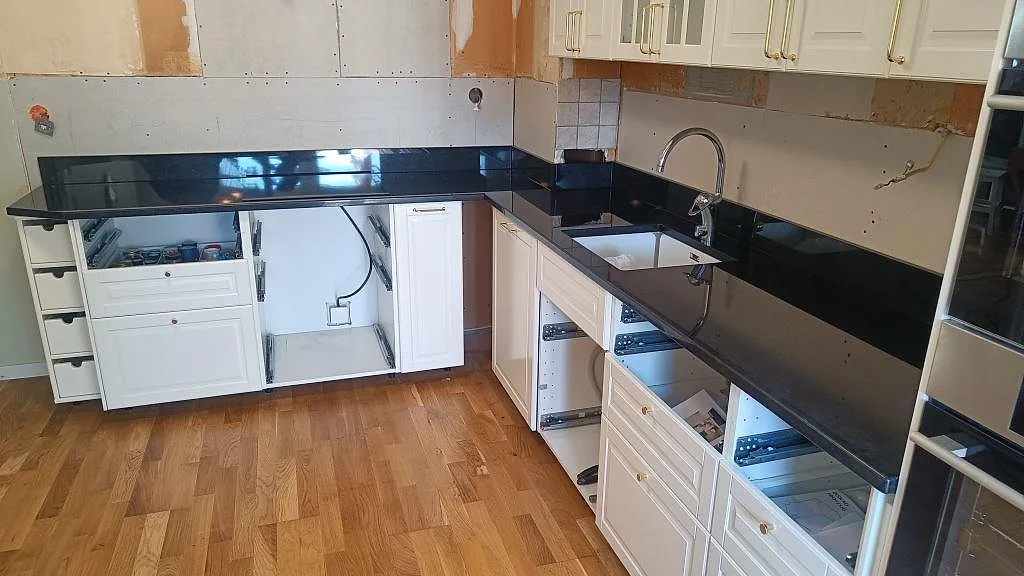 Køkkenbordplade i sort Nero Assoluto-granit, poleret, i L-form med underlimmad vask og vita köksluckor i ett køkken i Malmö.
