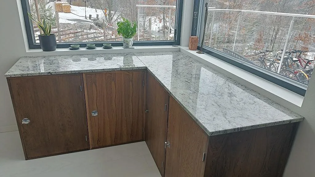 Køkkenbordplade i Antique White granit, poleret, L-formad og monteret på mørke träfronter med vindueskarme i ett køkken i Sandared.