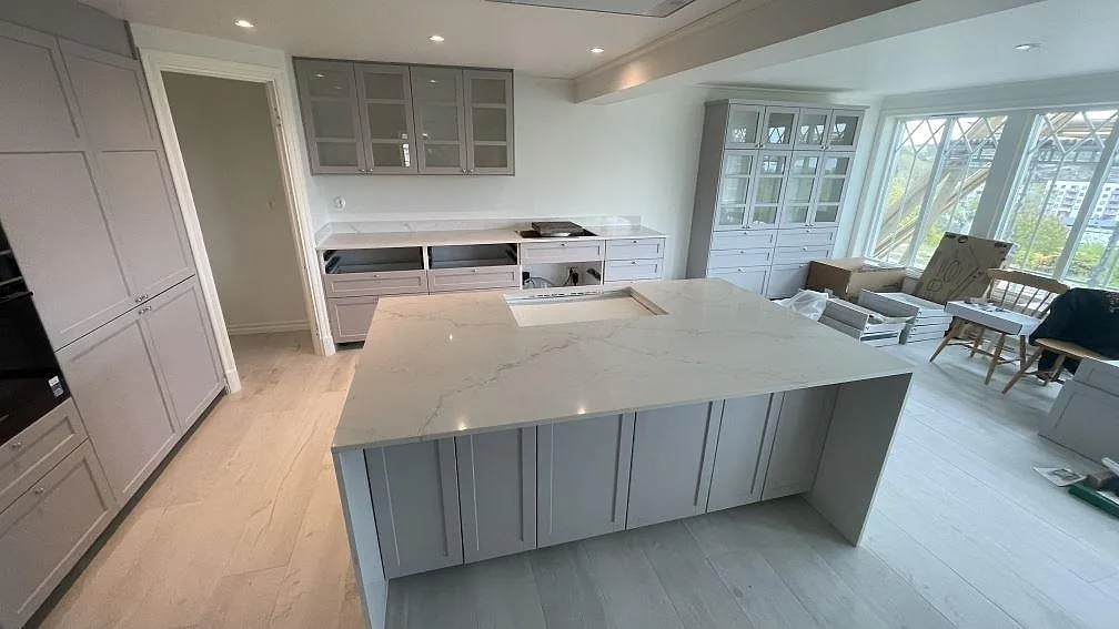 Køkkenbordplade og stor køkkenø i hvid Silestone Blanc Elysee, poleret, med grå åretegning og udskæring for vask i ett ljust køkken under montering i Strömstad.