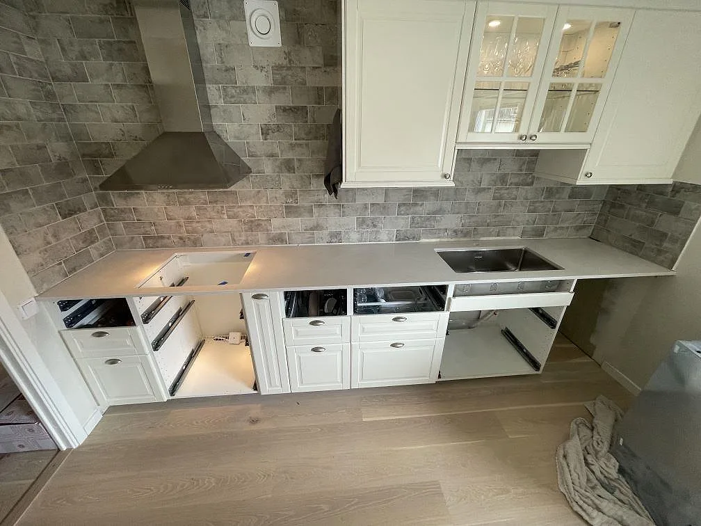 Køkkenbordplade i cremevit Silestone Blanco Maple 14, poleret 20mm, monteret under montering längs en L-formad køkkenbordplade mot grått marmormönstrat tegelkakel, med vita ramluckor og rostfri spiskåpa i ett køkken i Uppsala.
