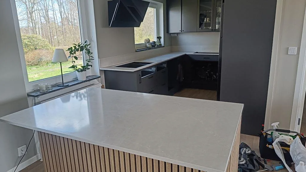 Køkkenbordplade og køkkenø i Silestone Desert Silver, poleret, i ett ljust køkken i Lidköping med integrerat stænkplade og mørke skabe under montering.