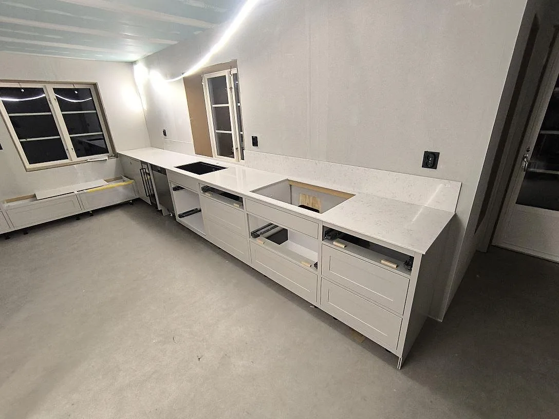 Lige køkkenbordplade i hvid Silestone Lagoon, poleret kvartskomposit, med uppvikt stænkplade, sort vask og hällurtag i ett køkken under montering i Avesta.