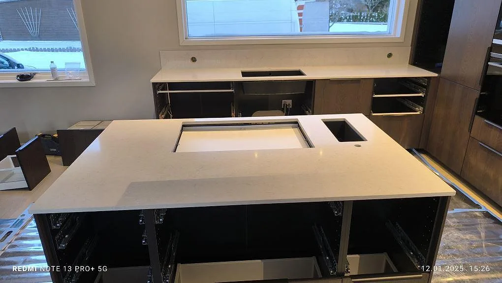Køkkenø og bänkrad i poleret Silestone Snowy Ibiza i ett køkken i Västervik, med lys kvartskomposit, förberedda udskæring og ett lavt stænkplade under fönstret.