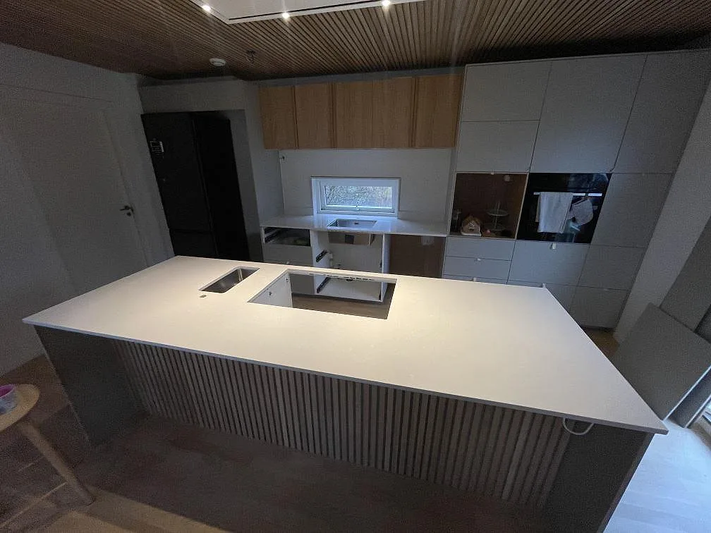 Køkkenbordplade i poleret Silestone Yukon, lys kvartskomposit, på en stor køkkenø med öppning for kogeplade og synlig rostfri vask i Tyresö.