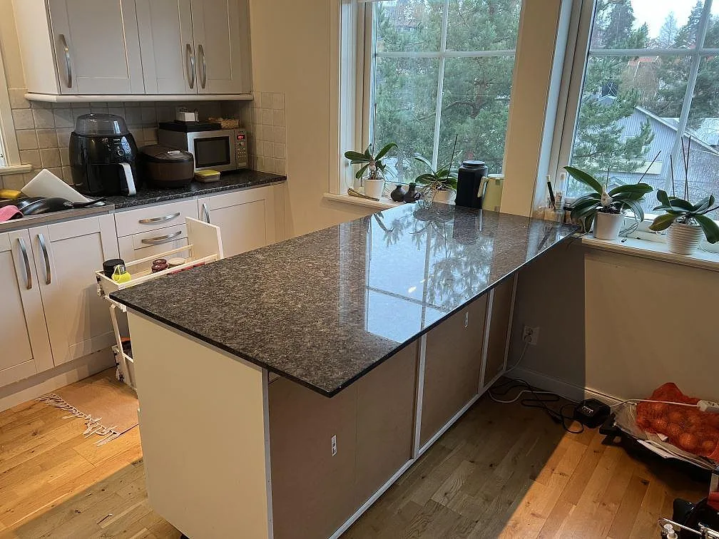 Køkkenø med 20 mm poleret Steel Grey granit i ett ljust køkken med stora vindue og växter i Saltsjö Boo.