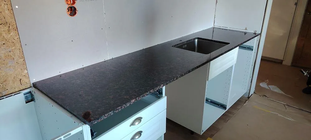 Køkkenbordplade i poleret Tan Brown-granit med diskhoöppning og mørk brun stenbild i ett køkken under montering i Björbo.