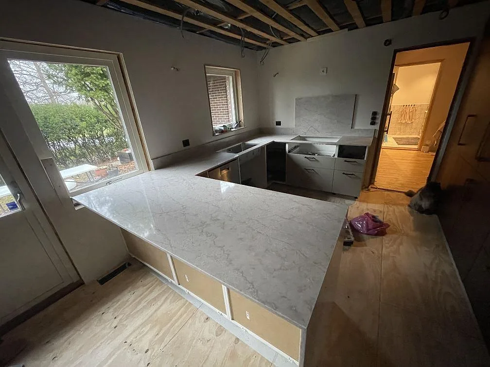 Køkkenbordplade i Technistone Glencoe, lys krämvit kvartskomposit med grå åretegning, poleret, monteret som halvö i ett U-format køkken under montering i Åkarp.