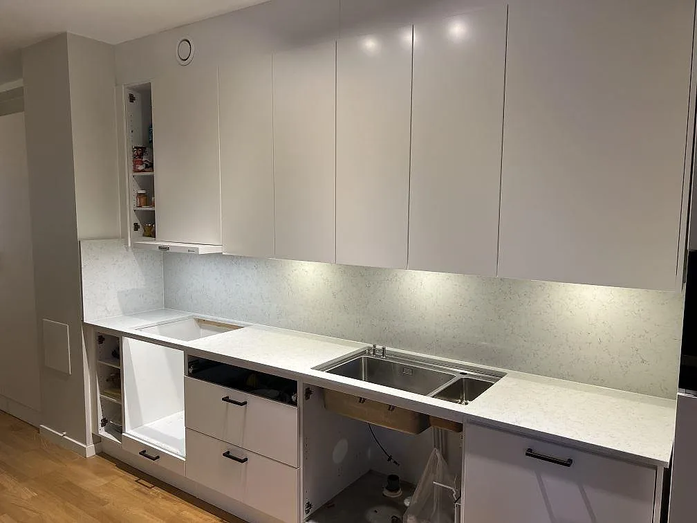 Køkkenbordplade i poleret Technistone Noble Carrara, lys kvartskomposit med grå åretegning, monteret längs væg i ett køkken i Sundbyberg.