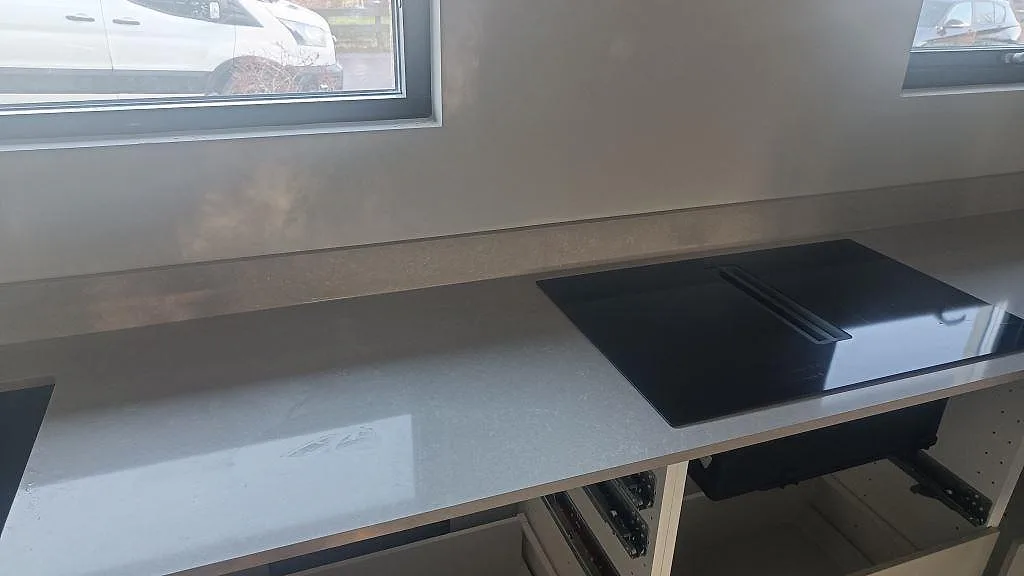 Køkkenbordplade i poleret Technistone Noble Ivory White, hvid kvartskomposit, med sort kogeplade og låg stänkkant i ett køkken i Kalmar.