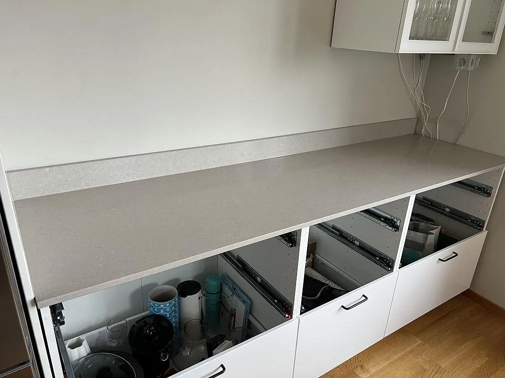 Lige køkkenbordplade i poleret Technistone Noble Ivory White, lys kvartskomposit med låg stänkkant ovanpå öppna vita köksstommar i Solna.