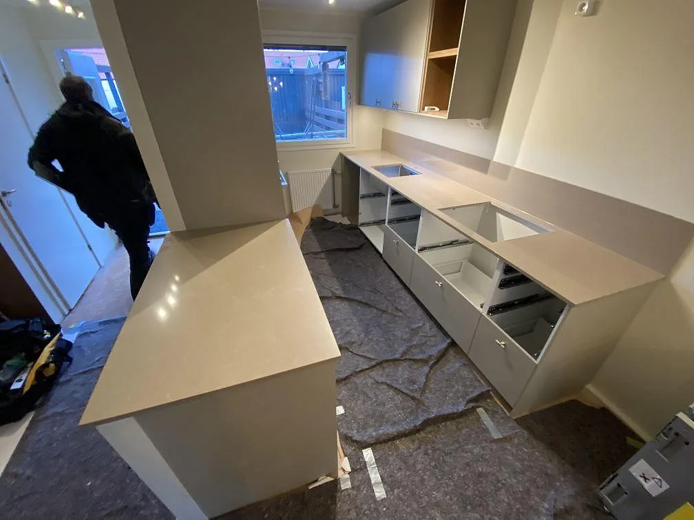 Køkkenbordplade i greige Technistone Noble Troya, poleret komposit, i ett L-format køkken i Tullinge med halvö, underlimmad vask og plats for induktionshäll under montering.