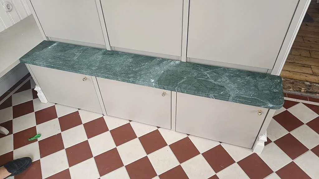 Marmorbordplade i grøn Verde Guatemala, mat 30 mm, på låg skåpsrad med lyse fronter og rödvitt rutgolv i Lilla Edet.