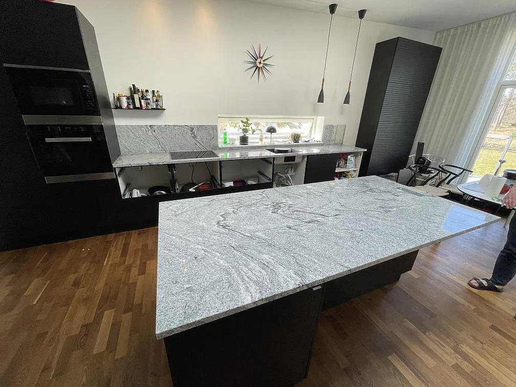 Køkkenbordplade og køkkenø i Viscont White granit, poleret med grå åretegning, i ett ljust køkken med mørke skabe og induktionshäll i Åhus.