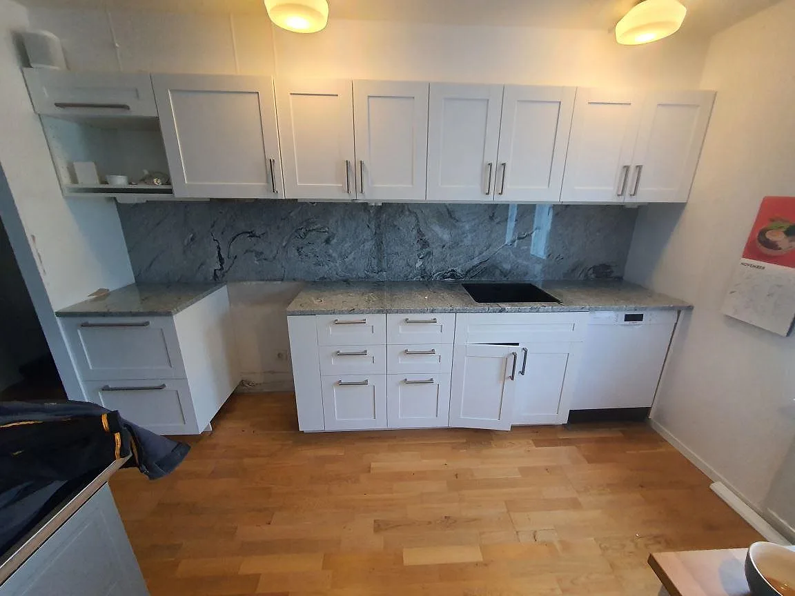 Køkkenbordplade i grå og hvid Viscont White granit, poleret, med heltäckande stænkplade i samme sten og mørk underlimmad vask i ett køkken i Bromma.