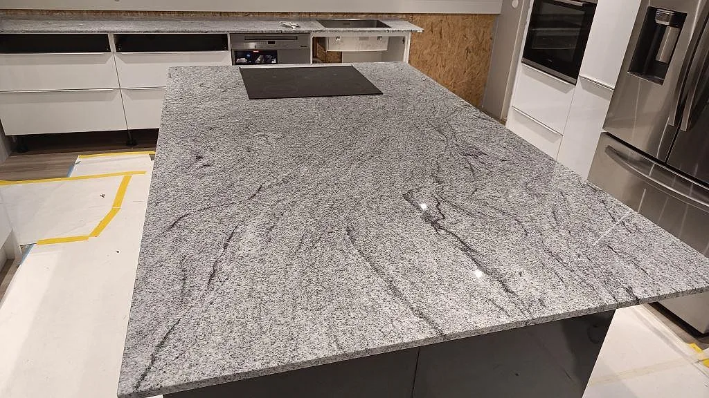 Stor køkkenø i poleret Viscont White granit med kogeplade og tydlig åretegning i ett køkken i Höllviken under montering.