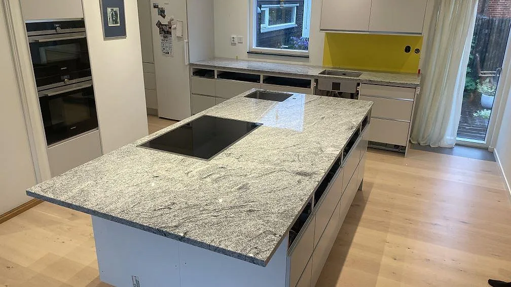 Køkkenø og Bordplader i Viscont White granit, 30 mm poleret, med sort kogeplade og ljusgrå åretegning i ett køkken i Malmö.