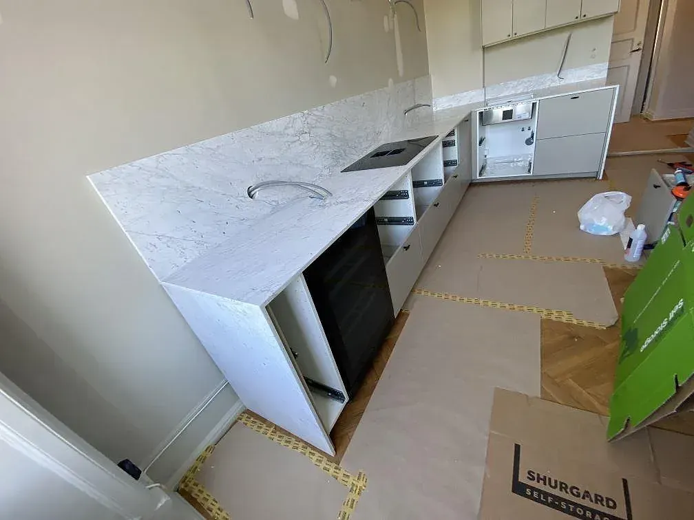 Køkkenbordplade i hvid Carrara C marmor, mat, längs en hel köksvägg med infälld induktionshäll og uppvikt stänkkant under montering i ett køkken i Malmö.
